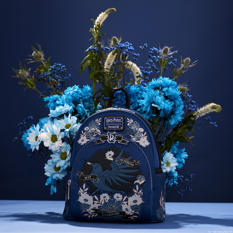 Blue Ravenclaw mini backpack featuring the Ravenclaw raven in the center of the front pocket surrounded by blue and white flowers, sitting against a blue background in front of a real bouquet of blue flowers 